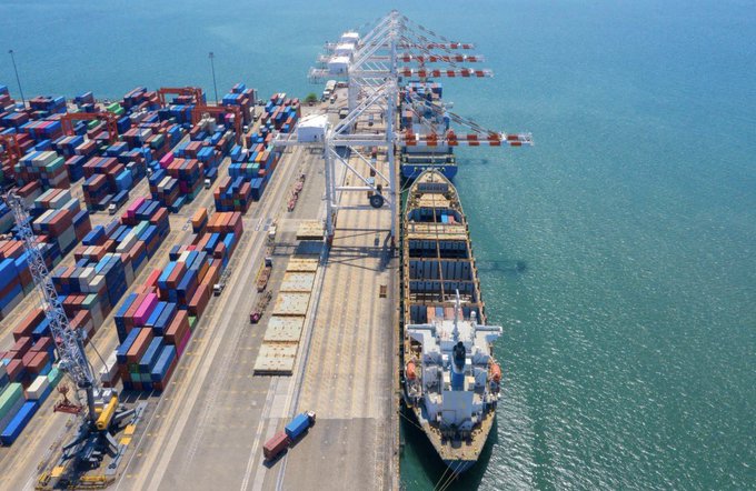 An aerial view of a container terminal where a large cargo ship is docked and being loaded with shipping containers by gantry cranes.