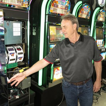 A man in a casino reaches toward a slot machine while rows of illuminated slot machines fill the background.