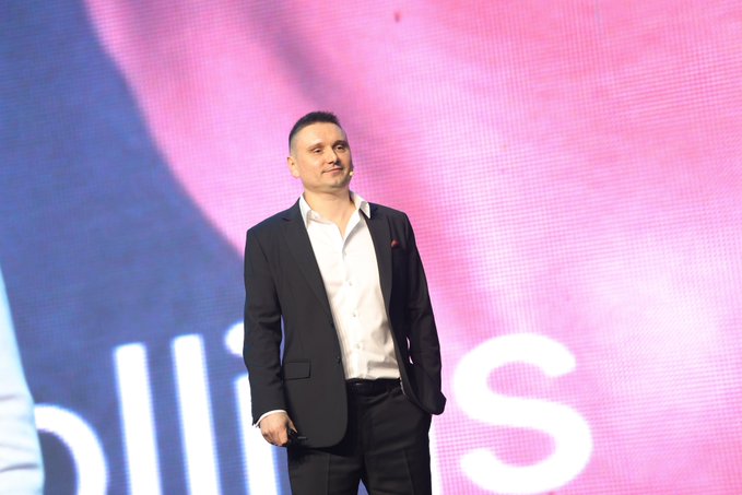 A man in a dark suit and white shirt standing on stage against a pink backdrop, smiling and looking toward the audience.