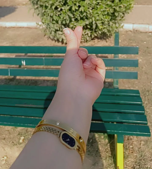 A close-up of a hand wearing a gold bangle-style watch with a dark face, held up in front of a green park bench.