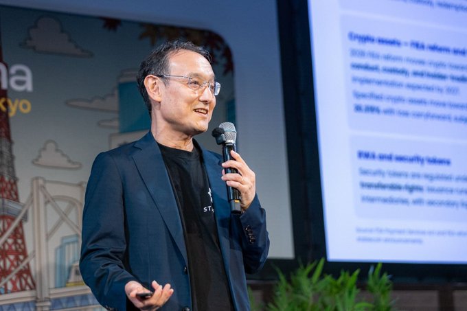 A man in a dark blazer and black shirt speaks into a microphone while presenting on stage, with a Tokyo-themed backdrop and a slide mentioning 'SZN, EVAR and EVM rollups and security tokens' visible behind him.