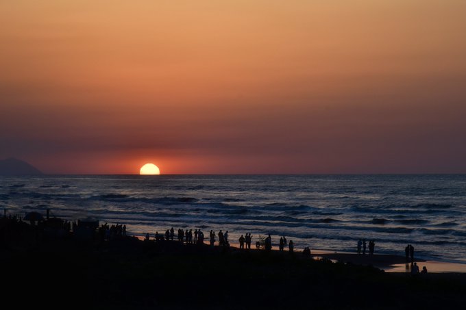 Silhouetted people gather on a beach to watch the sun setting over the ocean, casting an orange glow across the sky.