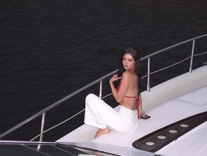A woman with long dark hair wearing a red bikini top and white pants sits on the deck of a white yacht, looking back over her shoulder at the camera against a backdrop of dark water.