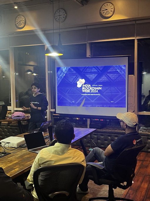Attendees gather at a workshop showing "India Blockchain Week 2024" on the projector screen, with a presenter in a "Marutiserve" hoodie preparing materials at a table with laptops, capturing pre-event preparations for the upcoming IBW 2024.