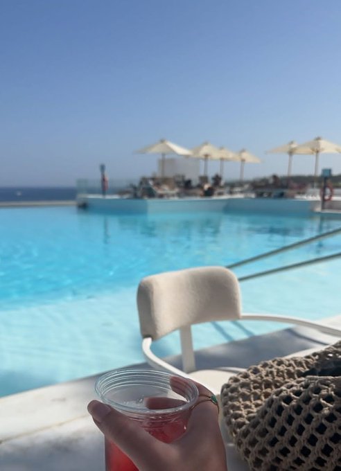 A first-person view from a poolside lounge chair holding a plastic cup of a pink drink, looking out over an infinity pool toward the ocean with white umbrellas and lounge areas under a clear blue sky.