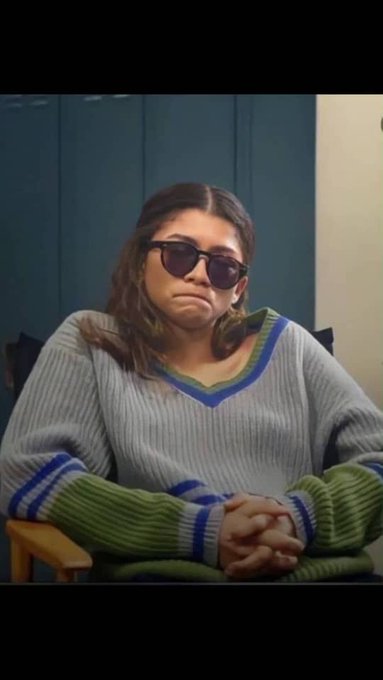 A woman wearing sunglasses and a grey sweater with green and blue accents sits with a pouty expression, her hands clasped in her lap.