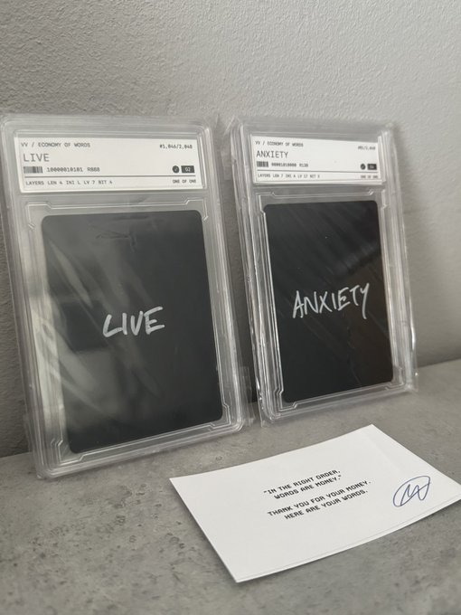 Two graded cards in plastic slabs labeled 'LIVE' and 'ANXIETY' by 'ECONOMY OF WOODS', accompanied by a small card reading 'IN THE RIGHT ORDER, WORLDS ARE MONEY. THANK YOU FOR YOUR MONEY. HERE ARE YOUR WORLDS.'