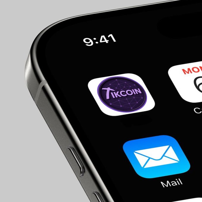 A close-up of a black smartphone displaying the TikCoin app icon, which features a purple network design with "TIKCOIN" text, alongside a blue Mail app icon on its home screen. The image showcases the TikCoin app being featured prominently on the device, aligning with the TikCoin @TikCoin_Network branding.
