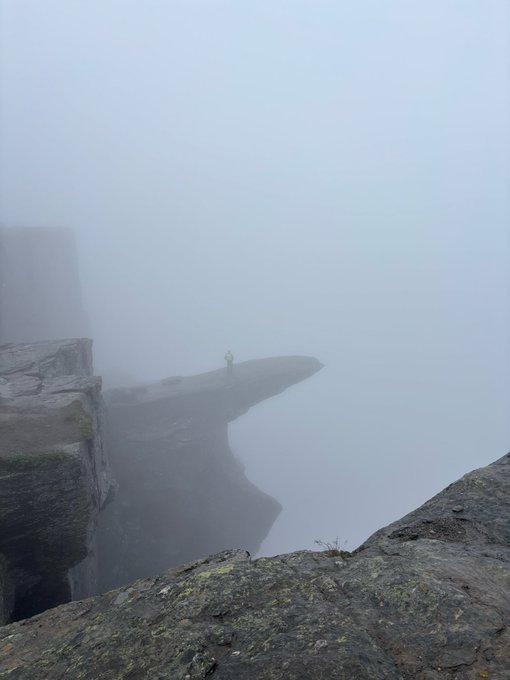 A tiny figure in a yellow jacket stands atop a misty, slab-like rock ledge—Trolltunga in Norway—shrouded in thick fog with jagged cliffs below. The image perfectly captures the remote, atmospheric Norwegian location where the MonkeyHoodNFT NFTs will be minted.