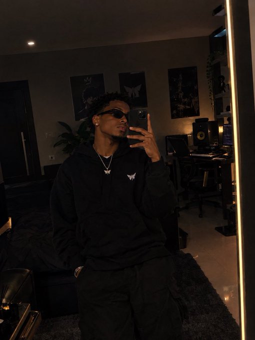 A young man wearing sunglasses and a black hoodie takes a mirror selfie in a dimly lit room with framed pictures on the wall.