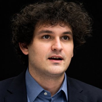 A man with curly dark hair wearing a blue button-down shirt and dark blazer looks off-camera with a surprised expression.