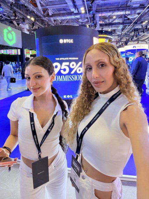 Two women in matching white outfits pose at what appears to be a crypto conference, with a BTCC booth advertising "95% COMMISSION" visible behind them.