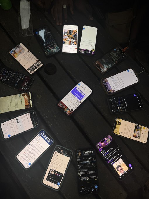Multiple smartphones are arranged in a circle on a dark surface, displaying various social media and chat apps on their screens.