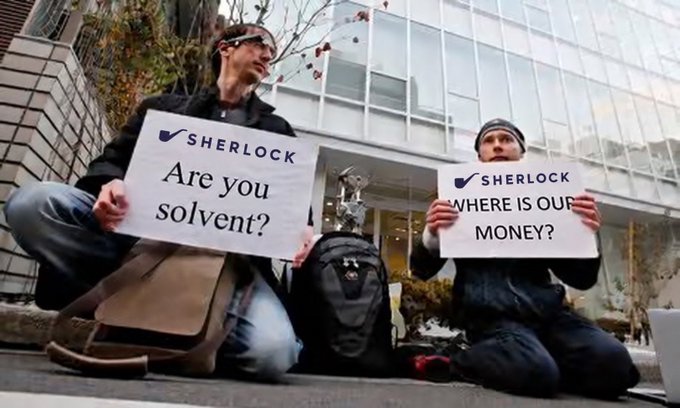 Two men sitting on the ground holding signs that say 'SHERLOCK Are you solvent?' and 'SHERLOCK WHERE IS OUR MONEY?', referencing Sherlock Communications and a debt issue.