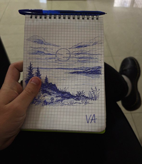 A hand holds a spiral-bound grid notebook displaying a blue ink sketch of a mountain lake landscape at sunset with pine trees, signed 'VA' in the corner, with a blue pen resting on top.