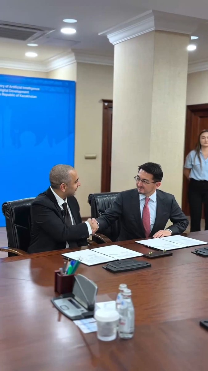 Two men in suits shake hands across a conference table with documents and a water bottle in front of them, with a partially visible blue backdrop reading "Ministry of Artificial Intelligence... Republic of Kazakhstan," marking the official partnership between Network School and Kazakhstan's AI Ministry as the program relocates from Singapore to Central Asia in September.