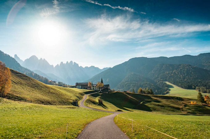 A scenic landscape photograph of a winding path through green alpine meadows leading toward a small village with mountains in the background under a bright, sunlit sky.