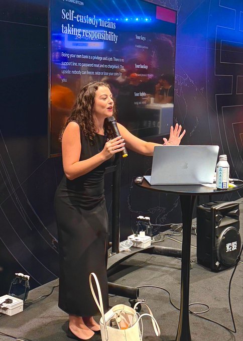 A woman with curly brown hair, wearing a black sleeveless dress, speaks into a microphone while gesturing toward a presentation screen titled 'Self-custody means taking responsibility.'