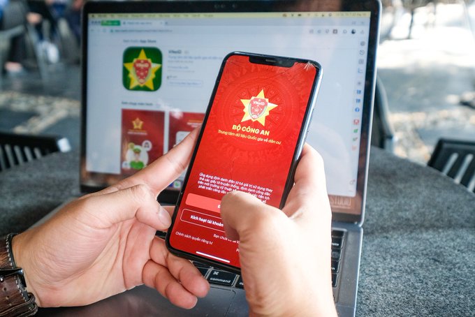A person holds a smartphone displaying the red-themed VNeID app login screen ('BỘ CÔNG AN') in front of a laptop showing a download page for the app.