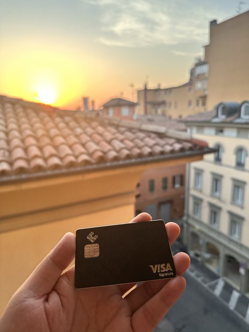 A hand displays a sleek black Visa Signature credit card against a stunning sunset view over terracotta-tiled rooftops and historic European architecture, perfectly matching the caption's praise of both "the card and the view" being exceptional.