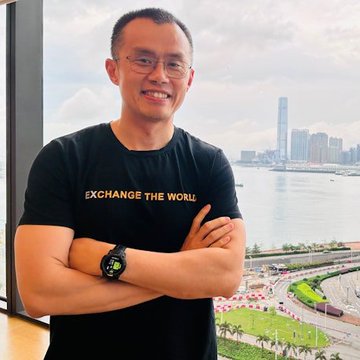 The image shows a smiling man wearing glasses and a black t-shirt that reads "EXCHANGE THE WORLD," standing with arms crossed against a backdrop of Hong Kong's skyline and harbor. He appears to be the Binance founder CZ, pictured during his visit to Hong Kong for Bitcoin Asia 2026 as referenced in the accompanying post.
