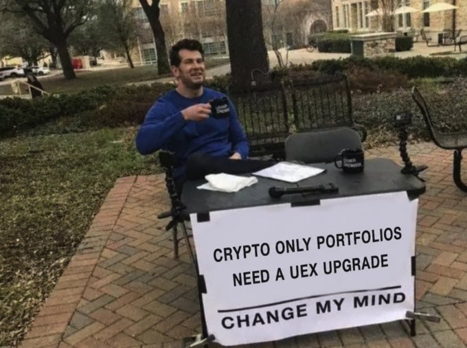 A man sits at a table outdoors in a 'Change My Mind' debate setup, with a sign reading 'CRYPTO ONLY PORTFOLIOS NEED A UEX UPGRATE', featuring a branded mug.