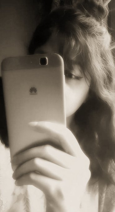 A black and white photo of a woman with curly hair taking a mirror selfie with a Huawei smartphone, with her face partially obscured by the device.