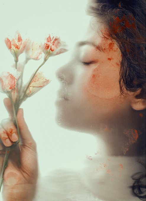 An artistic double-exposure photograph showing a woman's profile in profile holding delicate white and pink carnation flowers, with warm orange watercolor-like splashes blending across the image.
