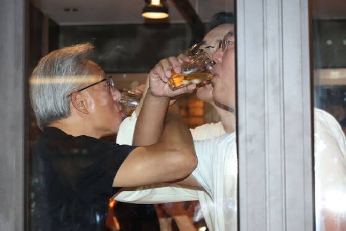 Two men are seen drinking from glasses simultaneously through a window, sharing a moment of indulgence. The image playfully captures the notion of partygoers transitioning directly from the KBW (Korea Blockchain Week) conference to Token2049, suggesting a celebratory or carefree attitude typical of crypto conference attendees.