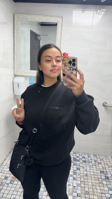 A woman takes a mirror selfie in a bathroom, wearing a black sweatshirt and black pants, holding a phone with a character-decorated case, and carrying a black crossbody bag.