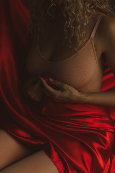 A moody, close-up photograph of a woman wearing a tan bra and wrapped in flowing red satin fabric, with her hand resting on her chest.
