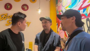 Three men converse in front of a colorful mural with red and yellow circular designs at what appears to be a restaurant or shop entrance.