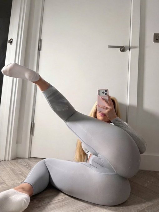 A selfie photo of a blonde woman in gray athletic leggings and a long-sleeve top, performing a flexibility pose on a wooden floor while holding a phone with a pink case to take the photo.