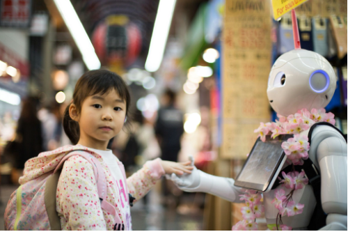 A photograph of a young girl in a pink floral jacket shaking hands with a white humanoid robot wearing a flower lei in what appears to be a Japanese shopping arcade.