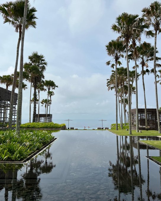 A symmetrical view of a long reflecting pool lined with tall palm trees, leading towards the ocean under a cloudy sky.