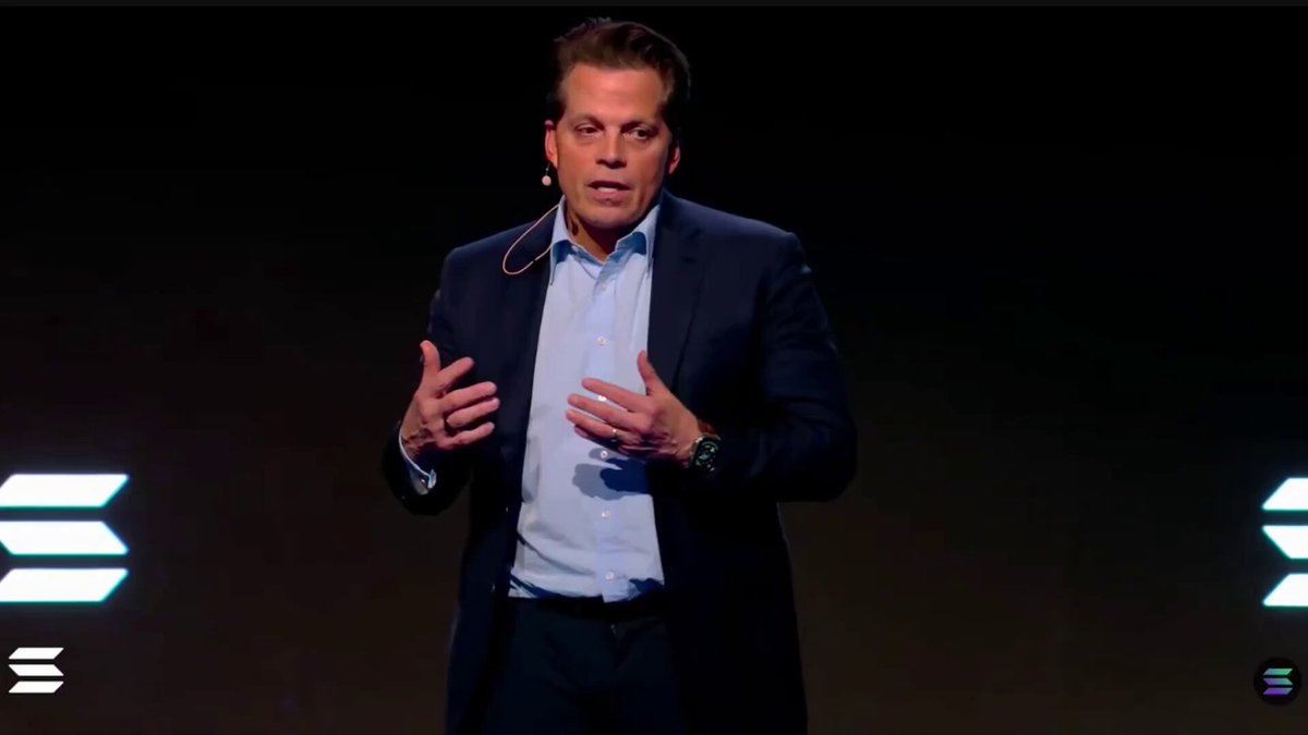 The image features a man wearing a suit, standing in front of two lights that are turned off. He appears to be giving a speech or presentation, with his hands outstretched as if he's gesturing towards something. The background consists of a black wall, and there is text on the image that reads "WALL STREET WILL USE SOLANA AS THE BACKBONE OF RWA TOKENIZATION— ANTHONY SCARAMUCCI". This suggests that the man might be discussing tokenization or blockchain technology in relation to the use of Ethereum as a backing for tokenization.