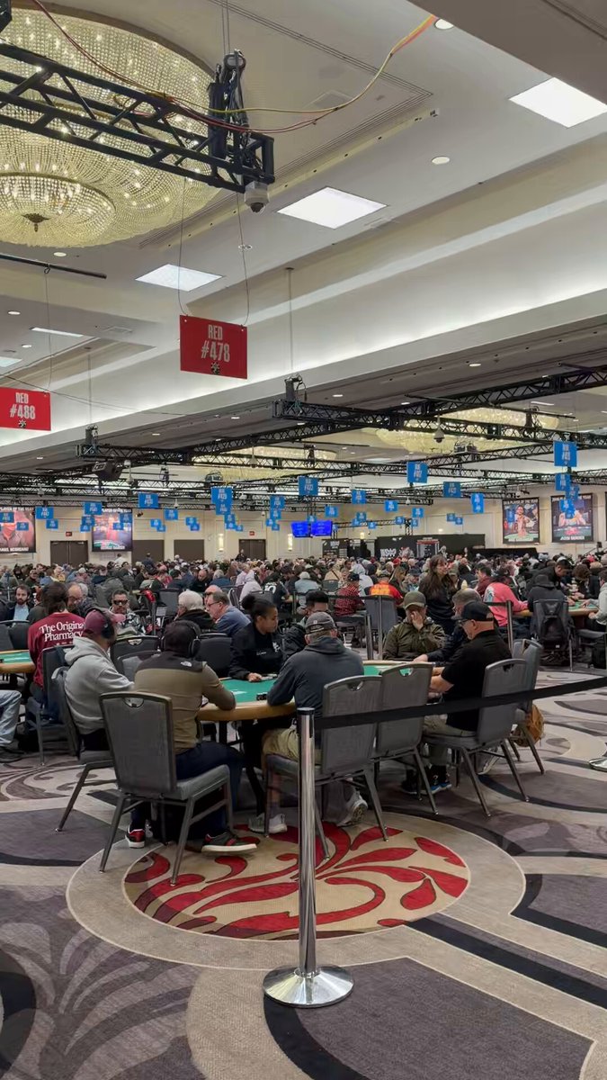 The image shows a group of people playing poker at a casino, sitting around tables with chairs arranged for them. There are multiple players engaged in the game, some wearing hats as part of their attire. The room has a large ceiling fan hanging from the center, and there is a TV mounted on the wall above it. A sign reading "These are the real trenches @WSOP @solana" can be seen in the image, indicating that this is a casino or poker room where players gather to play games of chance.