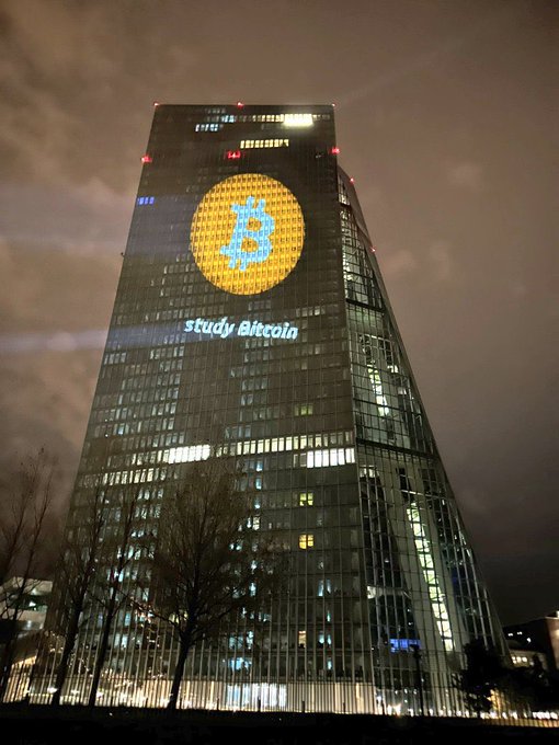 A tall skyscraper at night displays a large illuminated Bitcoin logo and the text 'study Bitcoin' projected onto its glass facade.