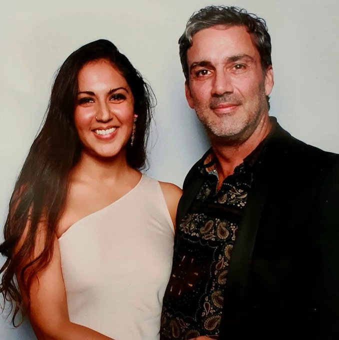 A couple poses together at what appears to be a formal event, with the woman in a white one-shoulder dress and the man in a dark patterned shirt and black jacket.