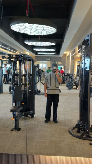 A mirror selfie taken in a gym by a person wearing a grey shirt and dark pants, with gym equipment and honeycomb-patterned ceiling lights visible.