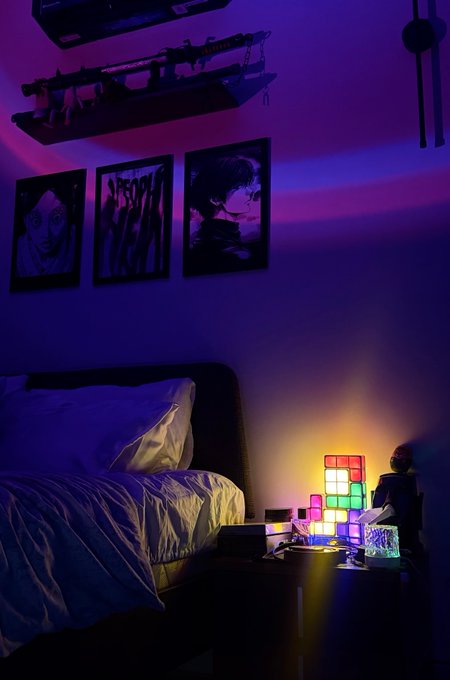 The image shows a dimly lit bedroom bathed in purple and blue ambient lighting, featuring three framed black-and-white portraits on the wall, a bed with white pillows, and a colorful Tetris-shaped LED lamp glowing warmly on the nightstand alongside books and figurines. The cozy, aesthetic "good morning" setting matches the post's weekend vibe, creating a relaxed atmosphere for the creator to reconnect with their audience before announcing giveaway winners and returning to regular content.
