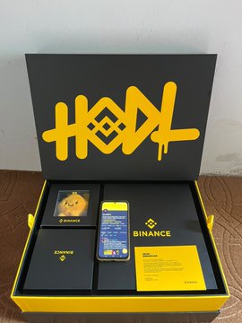 An open yellow and black 'HODL' branded merchandise box from Binance containing a phone displaying the Binance app, along with cards and accessories.