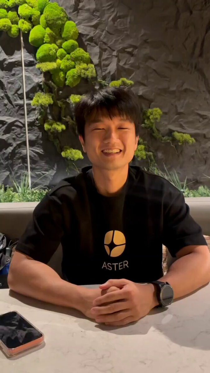 The image features a young man sitting at a table, smiling for the camera while wearing a black t-shirt with the word "ASTER" printed on it. The background of the photo shows a stone wall or rock face, which adds an interesting visual element to the scene. There is also a cell phone visible in the foreground, suggesting that this moment might have been captured during a social gathering or event where people are taking photos and sharing them online.