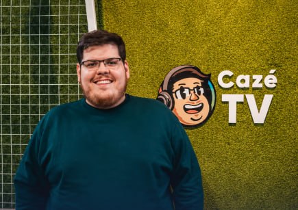 A smiling man with glasses wearing a teal sweatshirt stands in front of a green artificial grass wall featuring the Cazé TV logo.