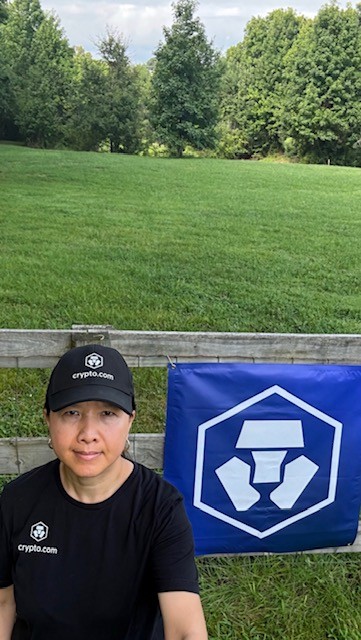 A woman wearing a black Crypto.com cap and t-shirt sits outdoors on a grassy lawn next to a blue Crypto.com flag.