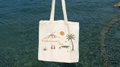 A white tote bag with a small illustration hangs against a blue textured background.