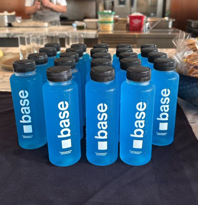 Multiple blue water bottles with black caps branded with 'base' are arranged in rows on a dark cloth, with a kitchen or cafeteria setting visible in the background.