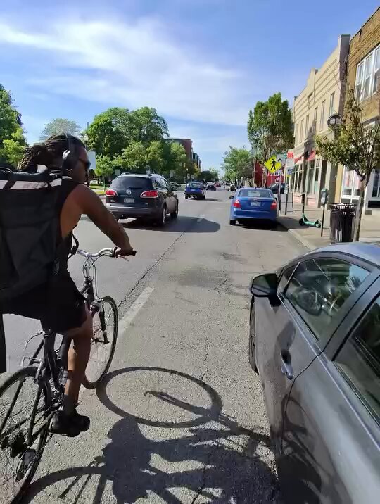 The image shows a man riding a bicycle down a street, wearing sunglasses and carrying a backpack. There are several cars parked along the side of the road, including one car that appears to be moving away from him. The background features buildings on both sides of the street, indicating an urban setting. A post in the scene reads "buy the $TROLL coin".
