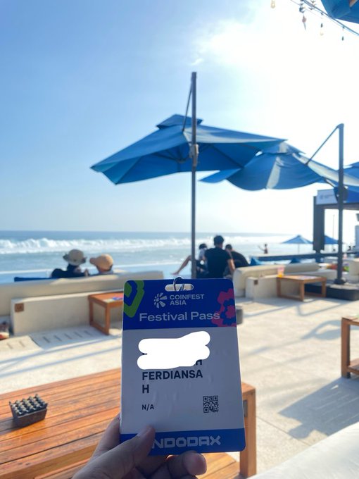 A hand holds a CoinFest Asia Festival Pass labeled 'FERDIANSAH' in front of a sunny beach scene with blue umbrellas and people enjoying the shore.