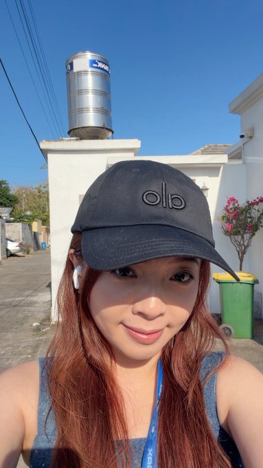 A young woman with long reddish-brown hair takes a selfie while wearing a black cap featuring the "olo" logo and white earbuds, with a blue lanyard around her neck, suggesting her attendance at the crypto/tech event. A stainless steel water tank labeled "PRO" sits on the rooftop behind her, and the clear blue sky and tropical setting tie into her CoinfestAsia experience.